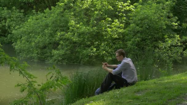 Επιχειρηματίας με κινητό τηλέφωνο που κάθεται στον πράσινο λόφο κρατώντας ένα Smartphone ξοδεύει το χρόνο σε πάρκο φρέσκα πράσινα χλόη δέντρα ηλιόλουστη μέρα του καλοκαιριού — Αρχείο Βίντεο