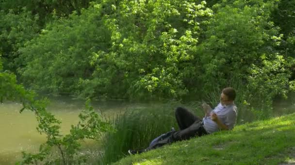 Geschäftsmann mit Handy auf grünem Hügel sitzend und Smartphone in der Hand verbringt Zeit im Park frisches grünes Gras Bäume sonnigen Sommertag — Stockvideo