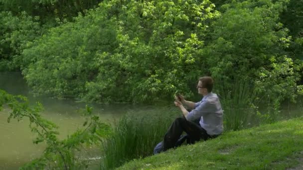 スマート フォンを保持緑丘の上に座って携帯電話を持ったビジネスマン公園新鮮な緑の草の木の日当たりの良い夏の日に時間を費やしています。 — ストック動画