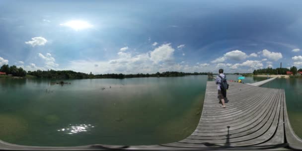 360vr Video Man stå på trä piren sjön solen lyser grön banken av floden Sea porlande vatten solen speglar turist går bort kusten Seaside sommardag — Stockvideo