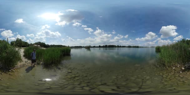 360vr Video Man Standing in Water op Lake Bank Turquoise transparant Water Reed bomen Man is kijken naar natuur wandelingen weg wolken zonnige zomer dag Blue Sky — Stockvideo