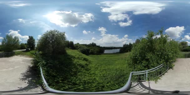 360vr βίντεο παρατήρηση Deck δίπλα στη λίμνη στο πάρκο πράσινα δέντρα ηλιόλουστη μέρα δρόμο δρομάκι πίσω από φράχτη δρόμο από πλακοστρώσεις πλακίδια μπλε ουρανό άσπρα σύννεφα ορίζοντα — Αρχείο Βίντεο