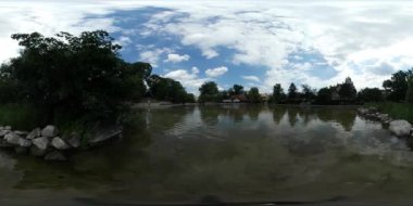 360vr Video adam Fording yapay göl Park yeşil ağaçlar bankada dekore ile taş binalar kırmızı çatılar dalgalanma gökyüzü yansıma bulutlu yaz günüdür