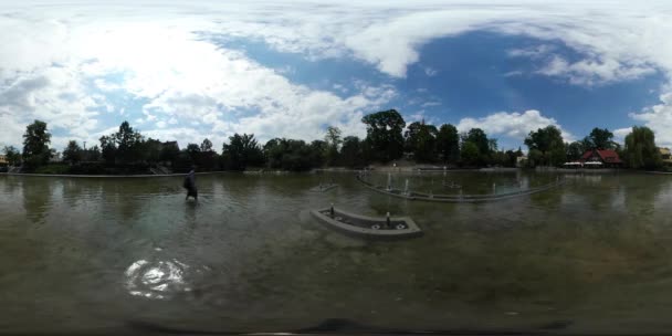 360vr video backpacker fording a lake fontänen in park green trees wellig sonnenreflexion in einem wasser gebäude auf einer bank backpacker tourist im sommertag — Stockvideo