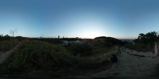 360Vr Vidéo Homme Marcher en pleine nature s'assoit et regarde la zone résidentielle à l'horizon Kiev Sol sablonneux Soirée d'été crépuscule Ciel bleu Vert Herbe — Video