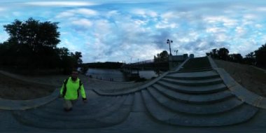 360vr Video adam tırmanma köprü Bisiklet Lane turist limon sarı ceketli için merdivenlerden yukarı geziniyor açık havada Dusk nehir bankalar bulutlu gökyüzü manzara olduğunu