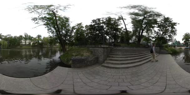 360vr 视频男人在公园路基铺设瓷砖楼梯新鲜绿树夏日天空云多云一天湖水荡漾的水休闲船附近 — 图库视频影像
