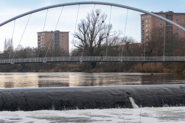 Mulde Nehri üzerindeki kemerli yaya köprüsünün gerçekçi bir fotoğrafı. Barajın çalkantılı suları ön planda görülürken, şehir binaları ve bulutlu bir gökyüzü altında çıplak ağaçlar arka planda yer alıyor..