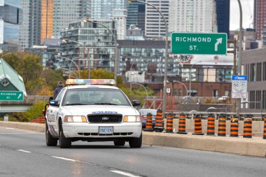 Toronto, ON, Kanada - 24 Ekim 2025: Işıkları açık beyaz bir polis kruvazörü arka planda şehir silüeti olan bir şehir otoyolunda ilerliyor.