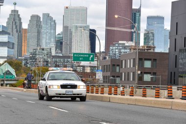Toronto, ON, Kanada - 24 Ekim 2025: Işıkları açık beyaz bir polis kruvazörü arka planda şehir silüeti olan bir şehir otoyolunda ilerliyor.