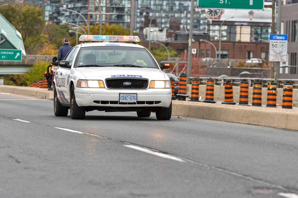 Toronto, ON, Kanada - 24 Ekim 2025: Işıkları açık beyaz bir polis kruvazörü arka planda şehir silüeti olan bir şehir otoyolunda ilerliyor.