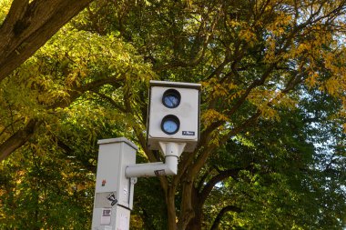 Toronto, ON, Kanada - 24 Ekim 2025: Beyaz otomatik hız kontrol kamerası (veya kırmızı ışık kamerası) yoğun yeşil ve sarı yapraklı sonbahar ağaçlarının olduğu bir kutba monte edilmiştir..