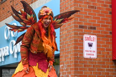 Toronto, ON, Kanada - 25 Ekim 2025 Yaprak kanatları ve renkli bir peruğu olan neşeli bir sonbahar perisi gibi giyinmiş, tuğla bir duvar ve güvenlik kamerası işaretinin yanında gülüyor.