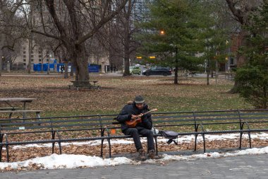 Toronto, ON, Kanada - 3 Aralık 2025: Parktaki bankta müzik aleti çalan adam, sonbahar yaprakları ve kar parçalarıyla çevrili, huzurlu bir anın tadını çıkarıyor