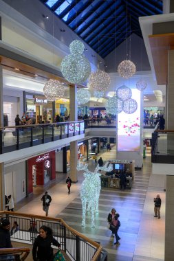 Toronto, ON, Kanada - 24 Aralık 2025: Alışverişçiler büyük bir ren geyiği ve asılı küreler de dahil olmak üzere aydınlatılmış Noel süslemeleriyle modern çok katlı bir alışveriş merkezinde yürüyorlar