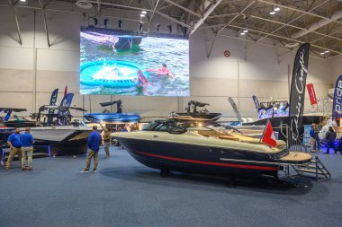 Toronto, ON, Kanada - 20 Ocak 2026: Su sporları gösteren bir video ekranı ile büyük bir kapalı sergi salonunda çeşitli sürat tekneleri ve deniz aksesuarları gezen ziyaretçiler