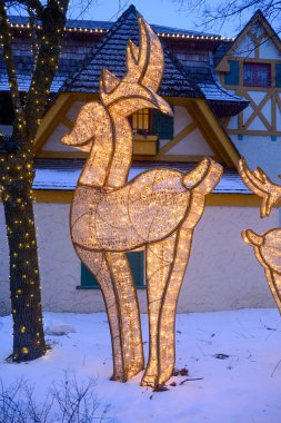 Toronto, ON, Kanada - 31 Aralık 2025: Aydınlatılmış ren geyiği heykeli kış festivali sırasında arka planda şenlikli bir bina ve yanan bir ağaç ile karda duruyor