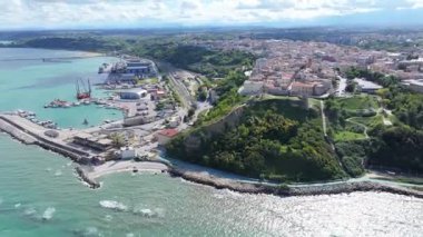 Ortona limanının hava manzarası turkuaz denizi, kıyıya yakın barışçıl dalgalar, demirlemiş tekneler, büyük gemiler ve parlak bir gökyüzü altında detaylı liman tesisleri yumuşak bulutlar ve sakin bir kıyı manzarası yaratıyor..