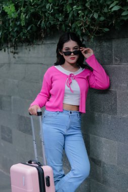 Elinde bavulu olan zarif bir kız tatile hazır, pembe bir hırka giyiyor, açık mavi kot pantolon ve şık güneş gözlüğü ile şehri keşfetmeye hazırlanıyor..