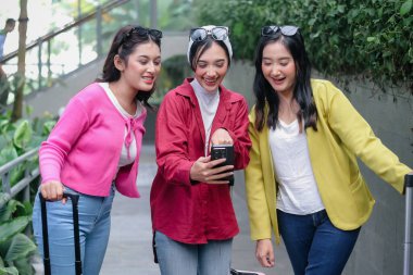 Üç genç, modern kadın birlikte seyahat ederken heyecanlı bir şekilde bir telefon ekranını paylaşıyorlar. Yanlarında bavullarıyla renkli giysiler içinde, mutluluk ve paylaşılan anların sevincini yayıyorlar..