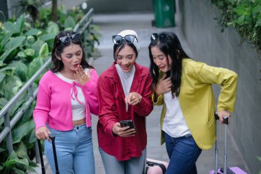 Üç genç Asyalı kadın, modaya uygun giyinmiş, bavul taşırken hevesli bir akıllı telefona danışıyorlar, neşe ve paylaşılan anlarla dolu bir yolculuğa hazırlanıyorlar..