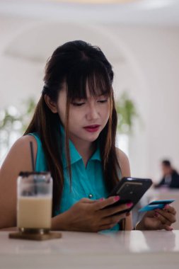 Bir kafede faturayı ödemek için cep telefonu kullanan genç bir kadın, internet üzerinden alışveriş yaparken elinde bir kredi kartı tutuyor..