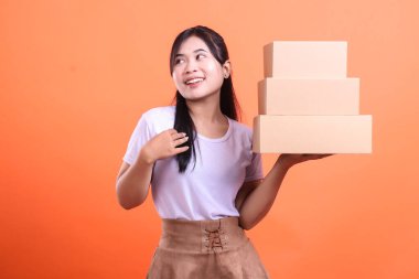 Mutlu bir Asyalı kadın elinde bir yığın karton kutu tutuyor. Yukarı bakıp gülümsüyor ve kutuları tutuyor. turuncu arkaplanda izole