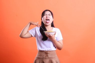 Genç bir kadın, bembeyaz bir kaseden yemek yedikten sonra, yüzü buruşmuş, turuncu arka planda izole bir şekilde yüzünü gösteriyor.