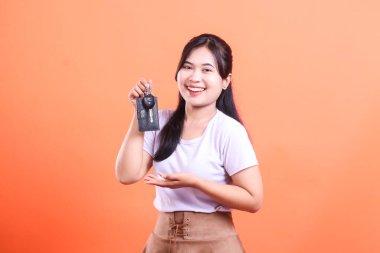 Gülümseyen Asyalı bir kadın araba anahtarı tutuyor ve açık bir avuç içi hareketiyle sunuyor. turuncu arkaplanda izole.