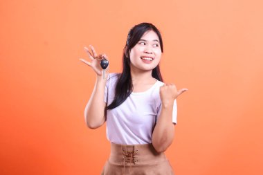 Mutlu bir Asyalı kadın arabanın anahtarlarını tutuyor ve gülümserken başparmağını yana çeviriyor. turuncu arkaplanda izole