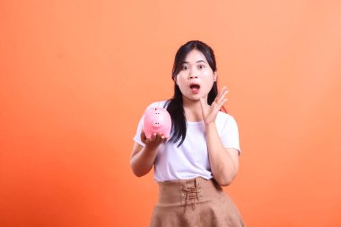 Asyalı bir kadın elinde pembe bir kumbara tutarken şok geçiriyor. turuncu arkaplanda izole.