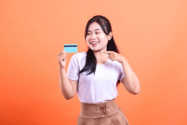 Gülümseyen bir kadın mavi bir kredi kartını işaret ediyor, memnun ve alışverişe hazır görünüyor, yeni finansal gücünü gösteriyor, turuncu arka planda izole edilmiş..