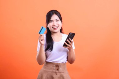 Elinde telefon ve kredi kartı olan gülümseyen genç bir kadın mutlu bir ifadeyle kameraya bakıyor. turuncu arkaplanda izole