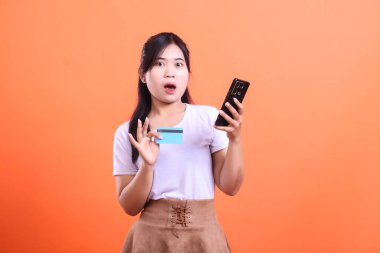 Şaşırmış genç bir kadın bir elinde kredi kartı, diğer elinde cep telefonu tutuyor. Gördüğü bir şey karşısında şok olmuş görünüyor. Turuncu arka planda izole edilmiş.