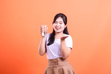 Neşeli genç bir kadın elinde bir bardak su tutuyor, susuzluğu ve sağlıklı yaşam tarzını teşvik ediyor, turuncu arka planda izole edilmiş.