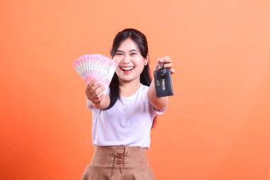 Neşeli bir tebessümle para ve araba anahtarı hayranı bir Asyalı kadın. turuncu arkaplanda izole.