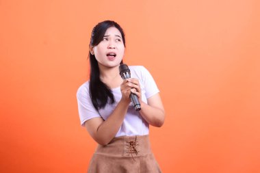 Genç Asyalı bir kadın mikrofonla şarkı söylüyor, ağzı açık bir şekilde turuncu arka planda izole bir şekilde yukarı bakıyor.