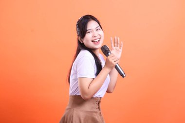 Genç, neşeli, mikrofonlu bir kadın izleyicilerine mutlu bir şekilde el sallıyor, yüzünde kocaman bir gülümsemeyle canlı ve heyecanlı görünüyor. turuncu arkaplanda izole