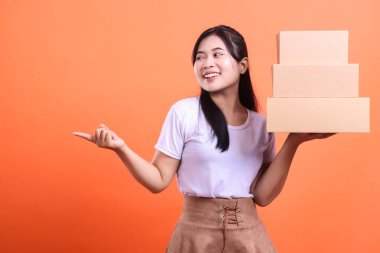 Neşeli bir yüzü olan genç bir kadın, bir elinde karton kutular tutarken diğer elinde, turuncu arka planda izole edilmiş bir yönü işaret ediyor..