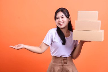 Neşeli genç bir kadın bir yığın karton kutu sunar. Teslimat yaptığı paketlerden memnun görünüyor ve elini uzatıyor, turuncu arka planda izole bir şekilde.