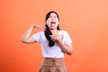 Genç bir kadın yiyeceğe karşı olumsuz bir tepki gösteriyor, bir kase erişte tutarken dilini dışarı çıkarıyor ve yüzünü buruşturuyor. turuncu arkaplanda izole