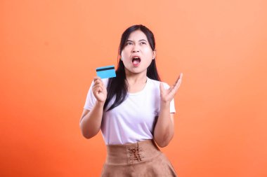 Elinde kredi kartı tutarken hayal kırıklığını ifade eden bir kadın, turuncu arka planda izole edilmiş para sorunlarıyla ilgili öfke ve mali stres gösteriyor.