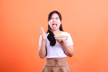 Elinde bir kase erişte tutarken surat asan genç bir kadın yediği yemekten tiksindiğini ve hoşnutsuzluğunu turuncu arka planda izole bir şekilde gösteriyor.