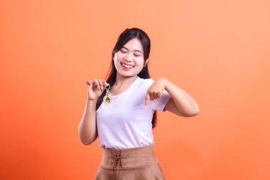 Mutlu genç bir kadın elinde evin anahtarlarını tutarken gülümsüyor, aşağı işaret ediyor, ev sahipliğini ve turuncu arka planda izole edilmiş yeni başlangıçları simgeliyor.