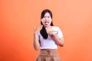 Genç bir kadın elinde bir kase erişte tutarken suratını asıyor, tatsız bir tada tepki veriyor, turuncu arka planda izole bir şekilde.