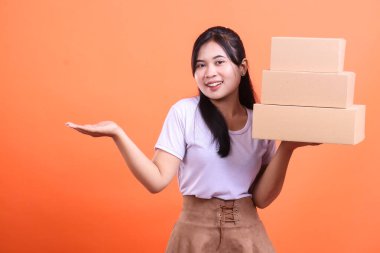 Mutlu bir Asyalı kadın bir elinde karton kutular tutar ve diğer elinde turuncu arka planda izole edilmiş bir şey gösterir.