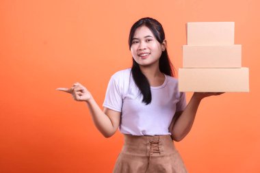 Mutlu genç bir kadın elinde bir yığın karton kutu tutuyor, bir eliyle sol tarafı işaret ediyor, bu da gösteriyor ya da gösteriyor. turuncu arkaplanda izole