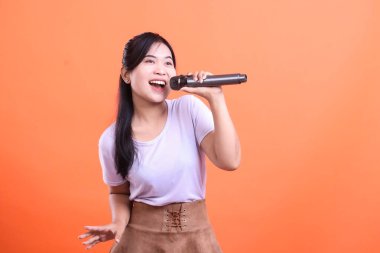 Heyecanlı genç Asyalı kadın mikrofona şarkı söylüyor, geniş bir gülümsemeyle, müziğe olan tutkusunu gösteriyor, turuncu arka planda izole edilmiş..