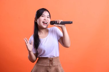 Genç bir kadın elini dinamik bir hareketle kaldırırken mikrofonla şarkı söylüyor. Turuncu arka planda izole edilmiş