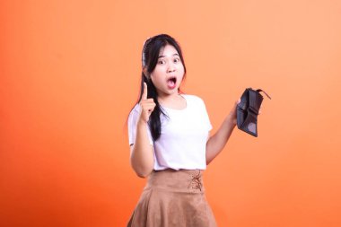 Şaşırmış görünen genç bir kadın boş bir cüzdan taşıyor. Beyaz bir tişört ve kahverengi bir etek giyiyor, turuncu arka planda izole edilmiş..